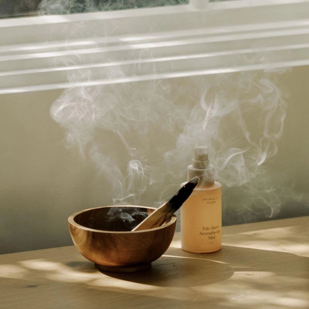 Wooden bowl with Primally Pure Palo Santo stick burning and a bottle of Primally Pure Palo Santo Aromatherapy Mist on a table surface. Primally Pure Palo Santo Collection of Non-Toxic Home Products