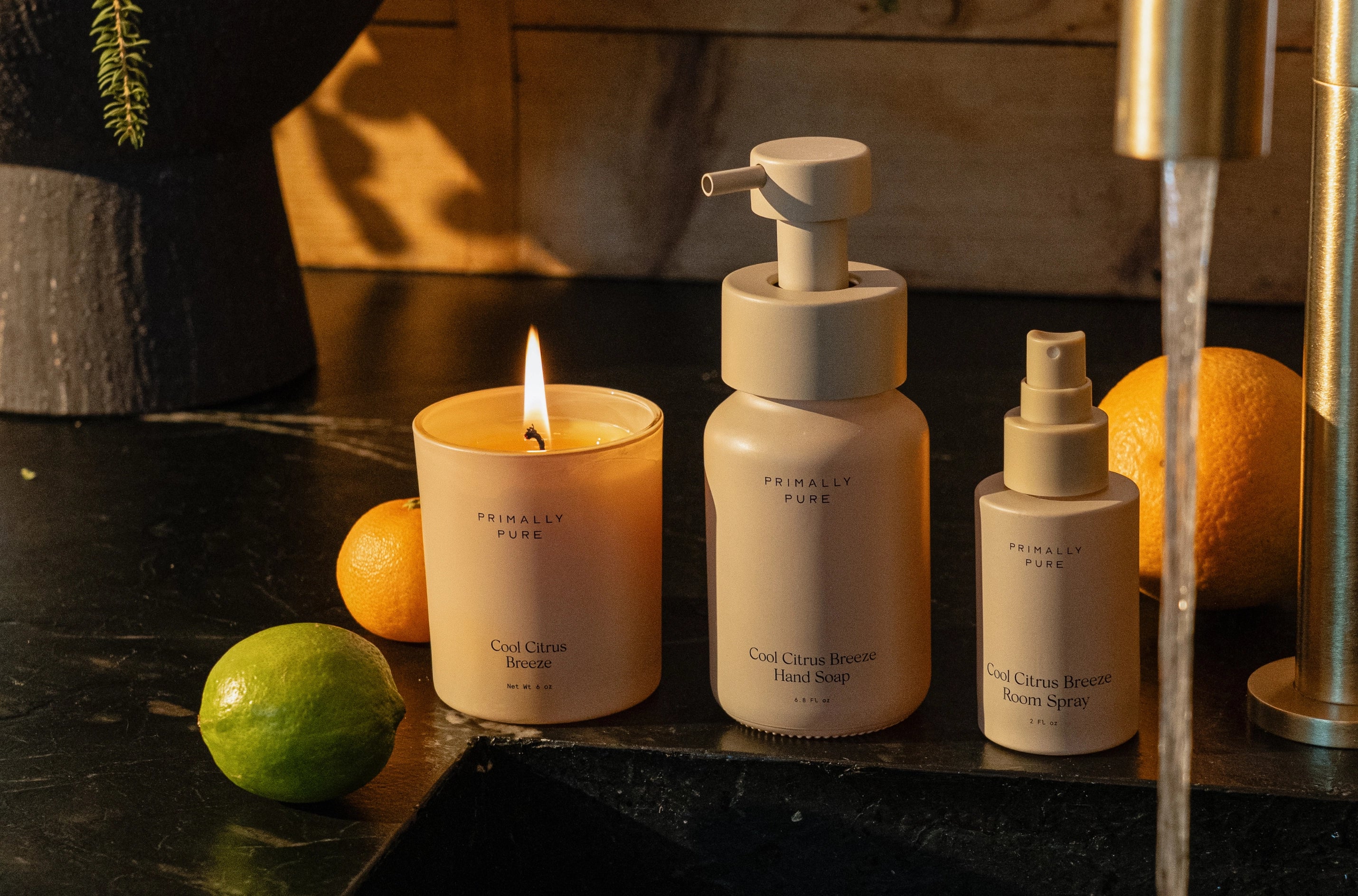 Three Primally Pure Cool Citrus Breeze Home Collection products on a dark surface with a lit candle and fruits.