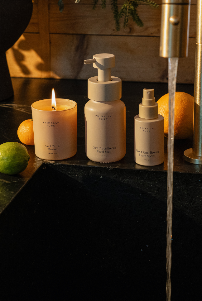 Three Primally Pure Home Collection products. A Hand Soap, Candle and Room Spray on a dark surface with a lit candle and fruit. Cool Citrus Breeze Non-Toxic Home Products by Primally Pure