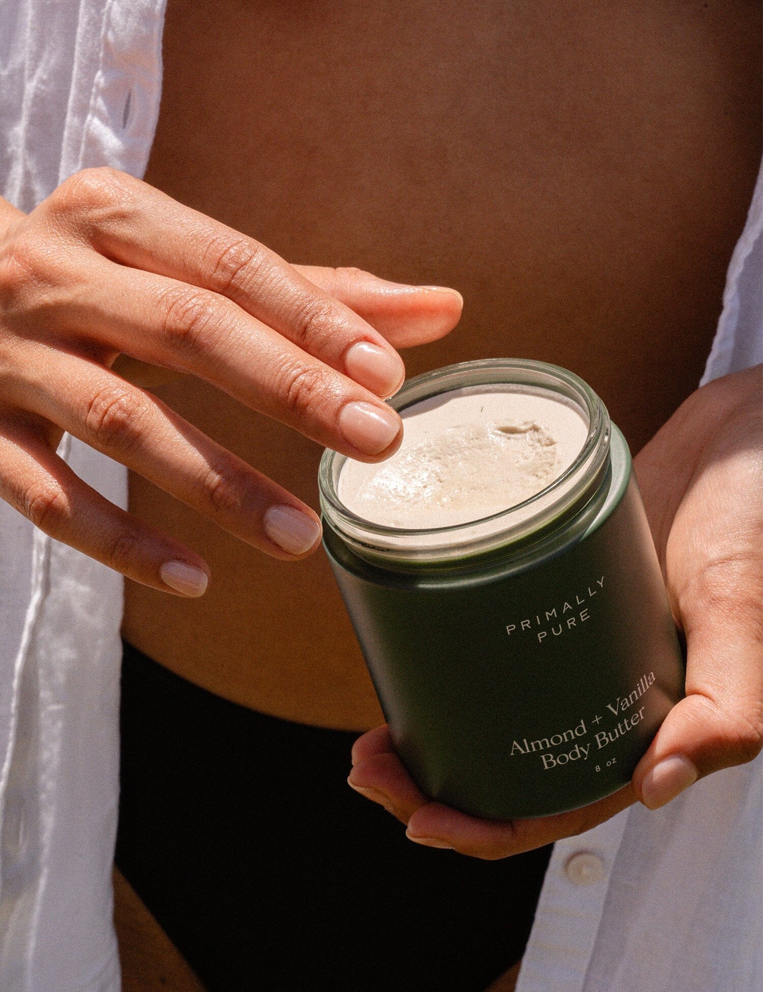 Person holding a jar of Primally Pure Almond + Vanilla Body Butter with fingers dipping into the creamy product, highlighting the texture and natural ingredients.