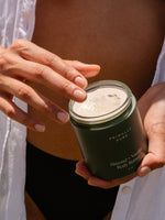 Person holding a jar of Primally Pure Almond + Vanilla Body Butter with fingers dipping into the creamy product, highlighting the texture and natural ingredients.