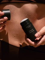 Close-up of a woman holding an open stick of Primally Pure Charcoal Deodorant near their midriff, with water droplets visible on their skin