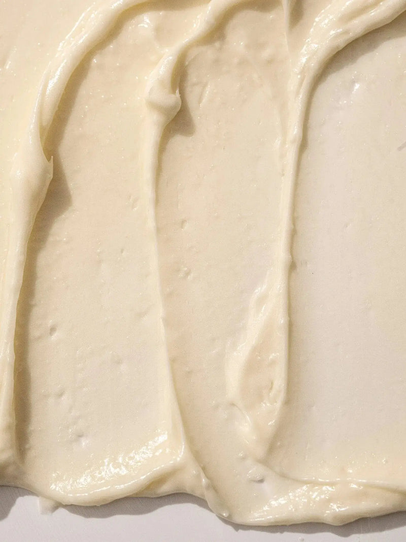 Person holding a green jar of Almond + Vanilla body butter using their other had to apply the body butter to their leg against a neutral background