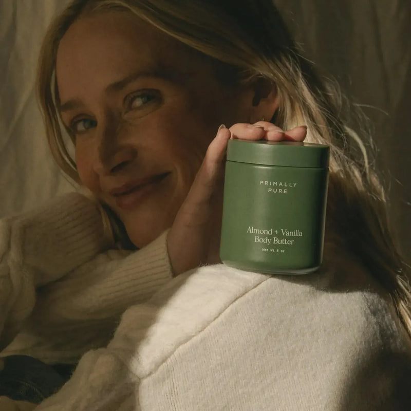 A woman in a cream knit sweater holds a green jar of Primally Pure Almond + Vanilla Body Butter against a warm, softly lit background.