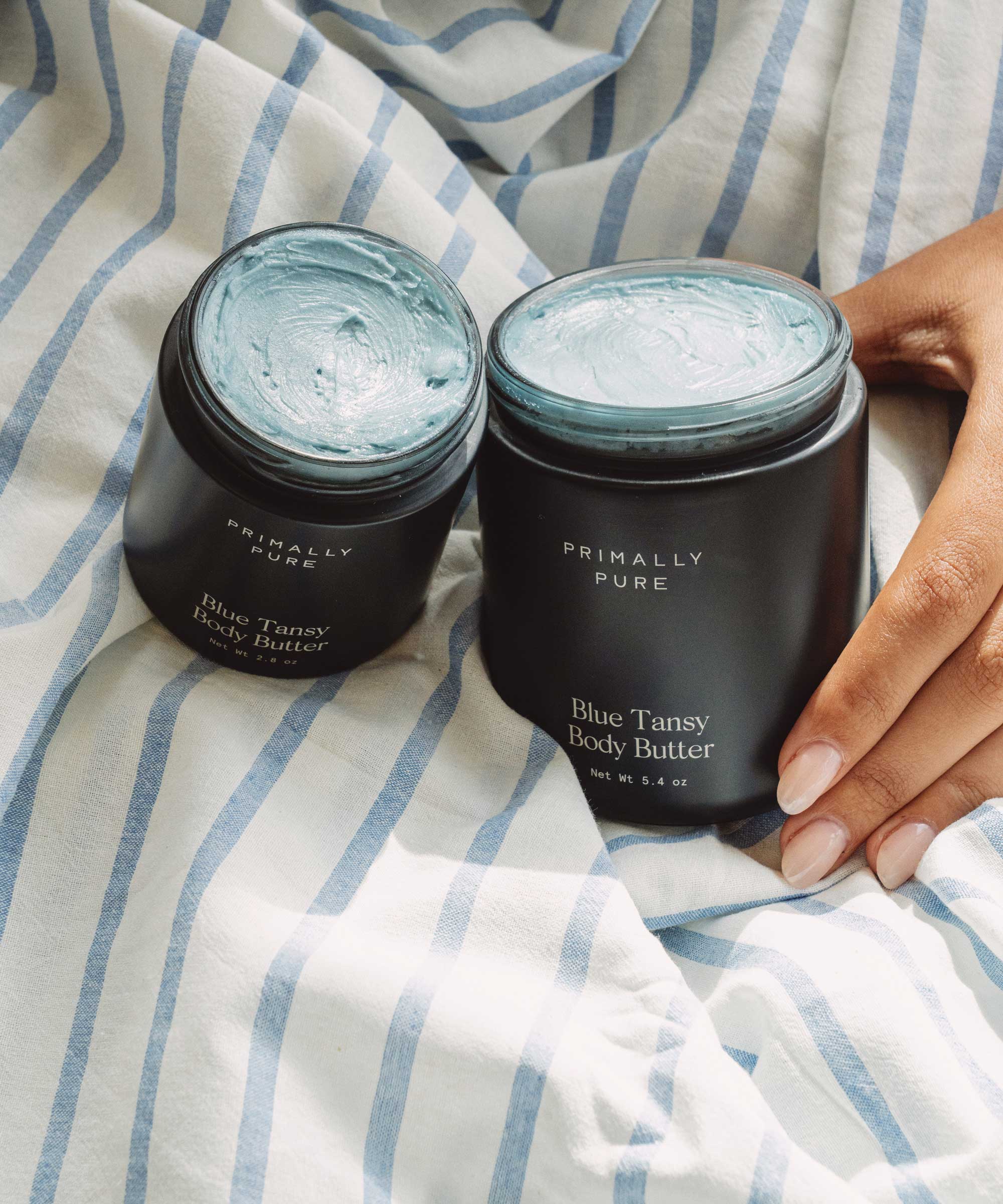 Two jars of Primally Pure Blue Tansy Body Butter on a striped fabric background.