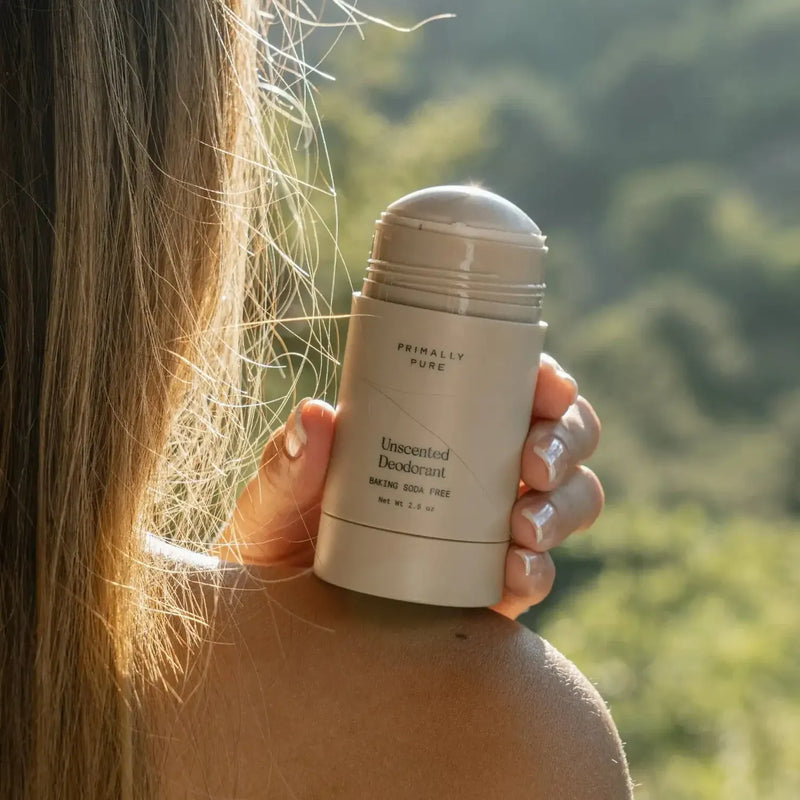A person outdoors holding a Primally Pure Unscented Deodorant Baking Soda Free stick against a blurred green natural background.