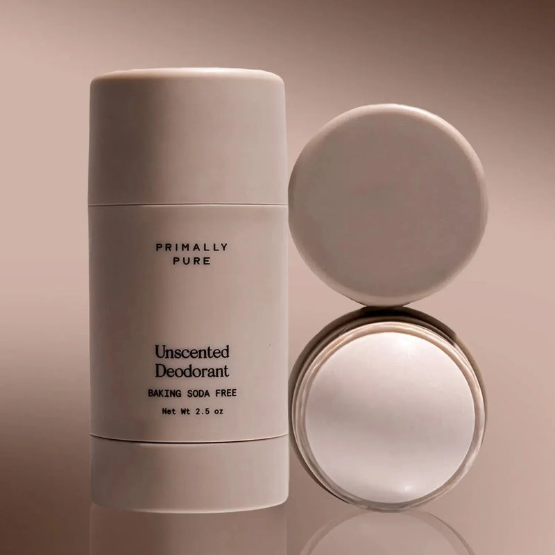 Primally Pure Unscented Baking Soda Free Deodorant stick in a matte white cylindrical container, shown alongside two circular white lids on a warm beige reflective surface.