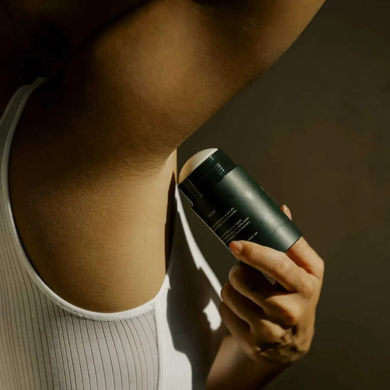 A close-up lifestyle shot of a person applying Bergamot + Eucalyptus deodorant stick from a white ribbed tank top against a dark brown background.