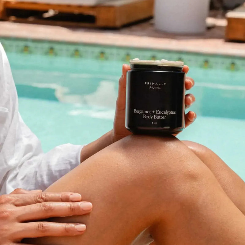 A person sitting by a swimming pool holds a black jar of Primally Pure Bergamot + Eucalyptus Body Butter. The person is wearing a white long-sleeve shirt, and the turquoise pool water is blurred in the background.