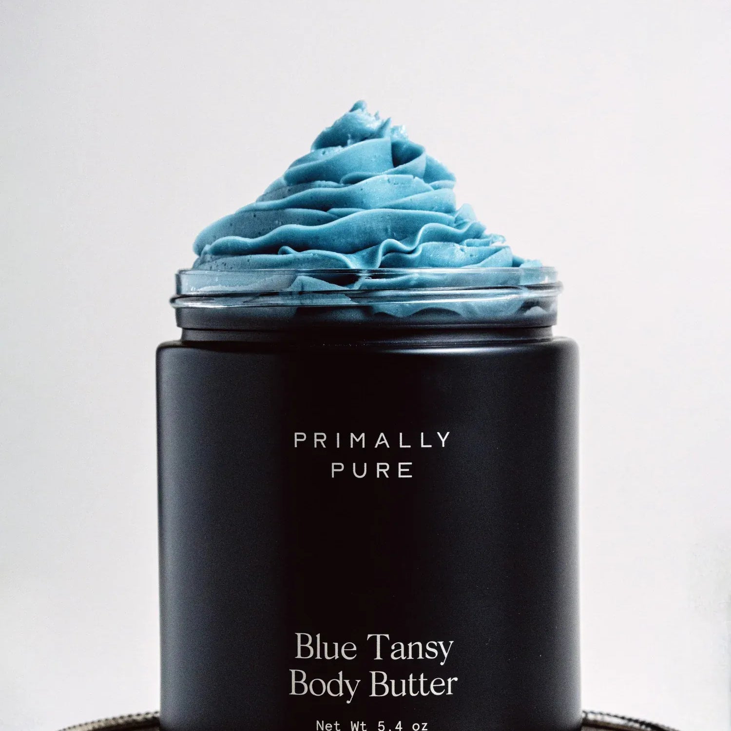 Black Jar of Primally Pure Blue Tansy on a white background showing the butter-like texture of Blue Tansy Body Butter
