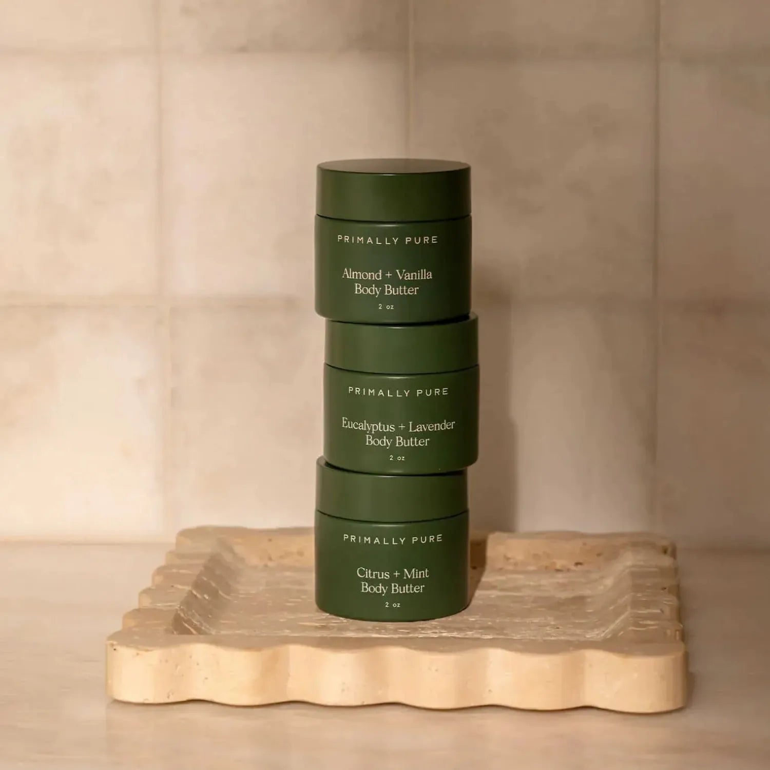 Three dark green jars of Primally Pure body butter stacked vertically on a scalloped beige stone tray against a neutral beige tiled background.