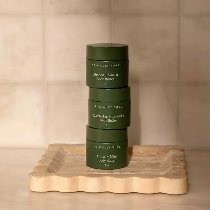 Three dark green jars of Primally Pure body butter stacked vertically on a scalloped beige stone tray against a neutral beige tiled background.
