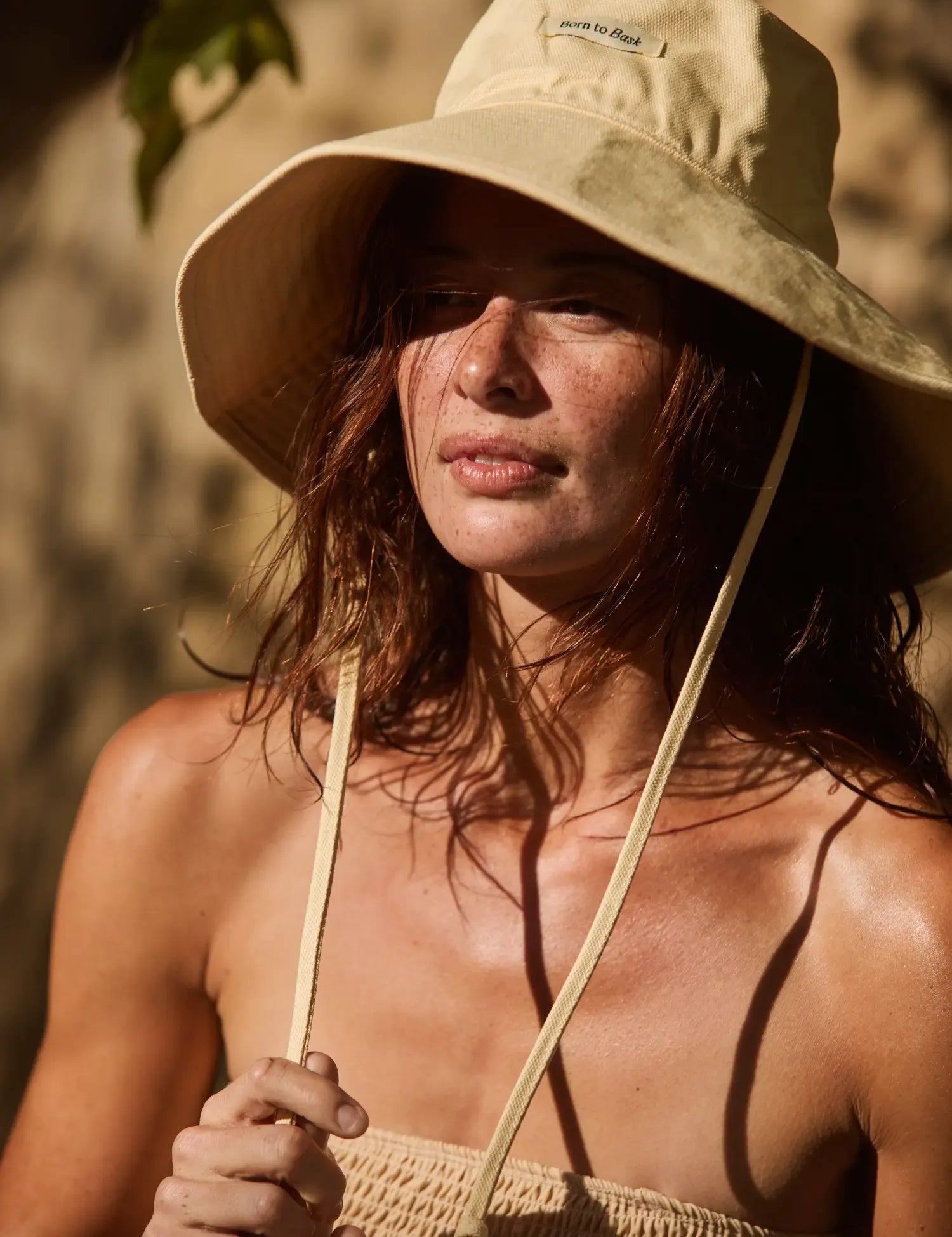 A woman wearing a beige wide-brim sun hat with a chin strap and a matching beige knit top, photographed in warm natural sunlight against a softly blurred outdoor background.