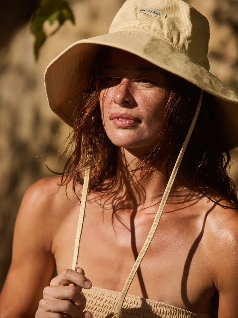 A woman wearing a beige wide-brim sun hat with a chin strap and a matching beige knit top, photographed in warm natural sunlight against a softly blurred outdoor background.