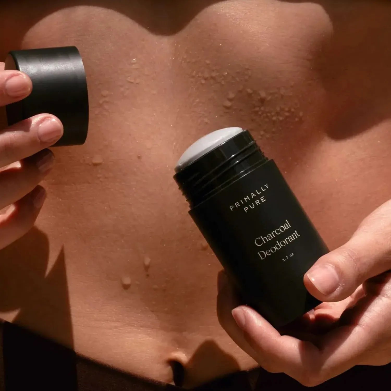 Close-up of a woman holding an open stick of Primally Pure Charcoal Deodorant near their midriff, with water droplets visible on their skin