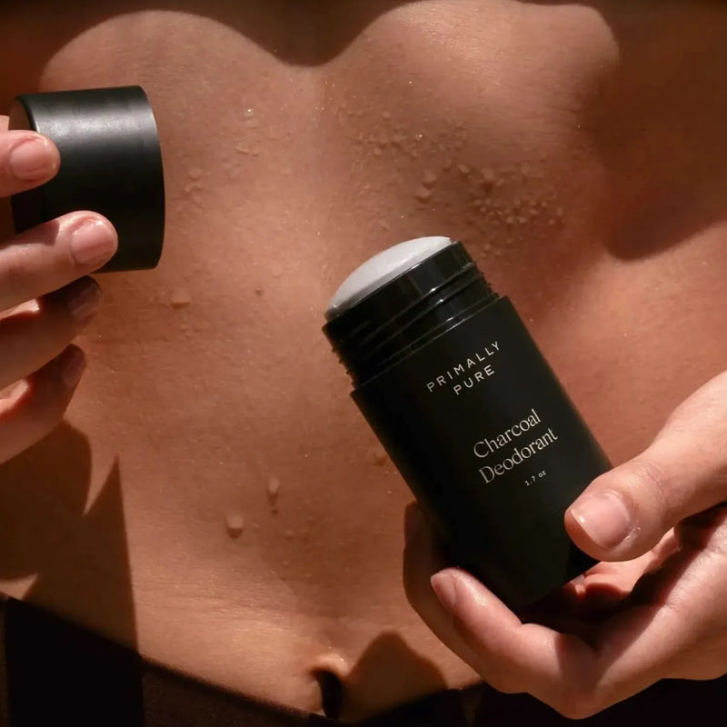 Close-up of a woman holding an open stick of Primally Pure Charcoal Deodorant near their midriff, with water droplets visible on their skin