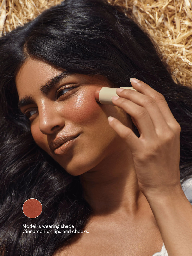 Woman applying Primally Pure Lip + Cheek Tint  with a focus on her face, using a shade labeled Cinnamon.