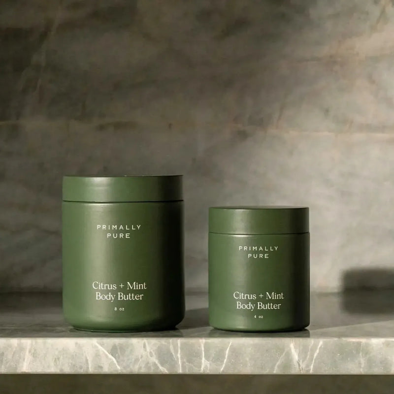 Two Primally Pure Citrus + Mint Body Butter jars in matte olive green, one large and one medium, displayed on a gray marble surface against a textured gray stone wall.