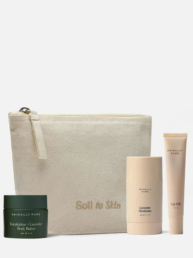 A skincare set from Primally Pure on a light gray background, featuring a beige canvas pouch with 'Soil to Skin' printed on it, a dark green eucalyptus + lavender body butter jar, a beige lavender deodorant stick, and a beige lip oil tube.