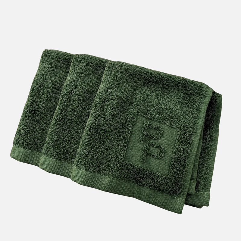 Three folded dark green Primally Pure Cleansing Clothes with ribbed borders and an embroidered monogram logo, arranged diagonally on a light gray background.