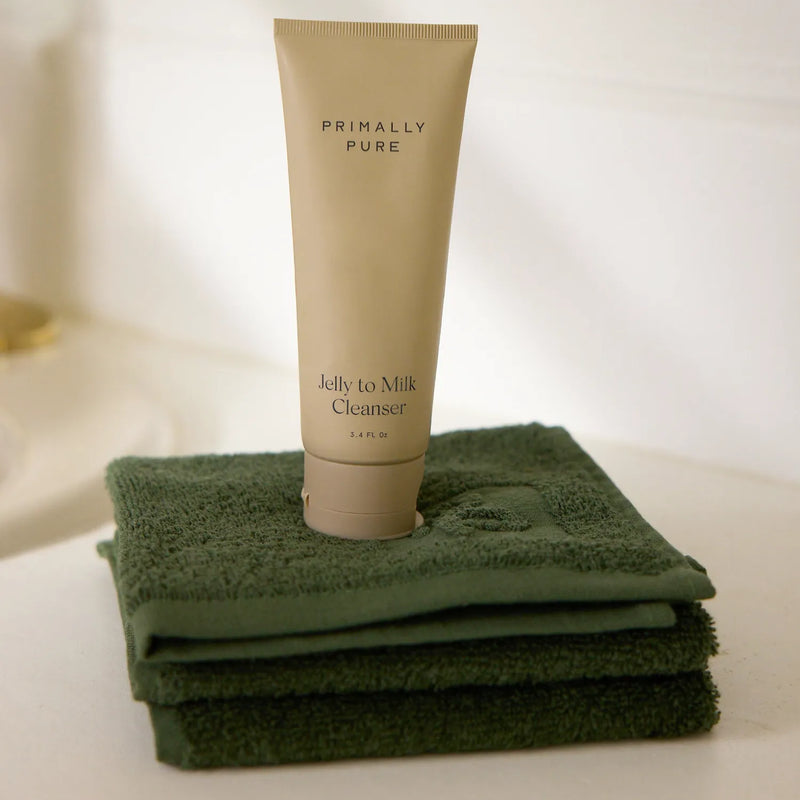 Primally Pure Jelly to Milk Cleanser tube standing upright on a stack of folded dark green Cleaning Cloth 3-Pack on a light surface.