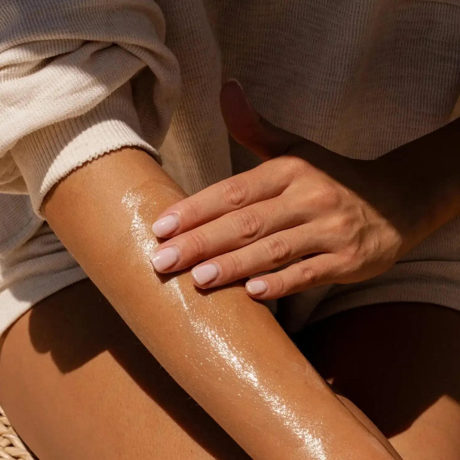 Close-up of a person's forearm and hand with a glossy, shiny moisturizing or cosmetic cream being rubbed in. The person is wearing a beige ribbed knit sweater, and the warm sunlight casts soft shadows.