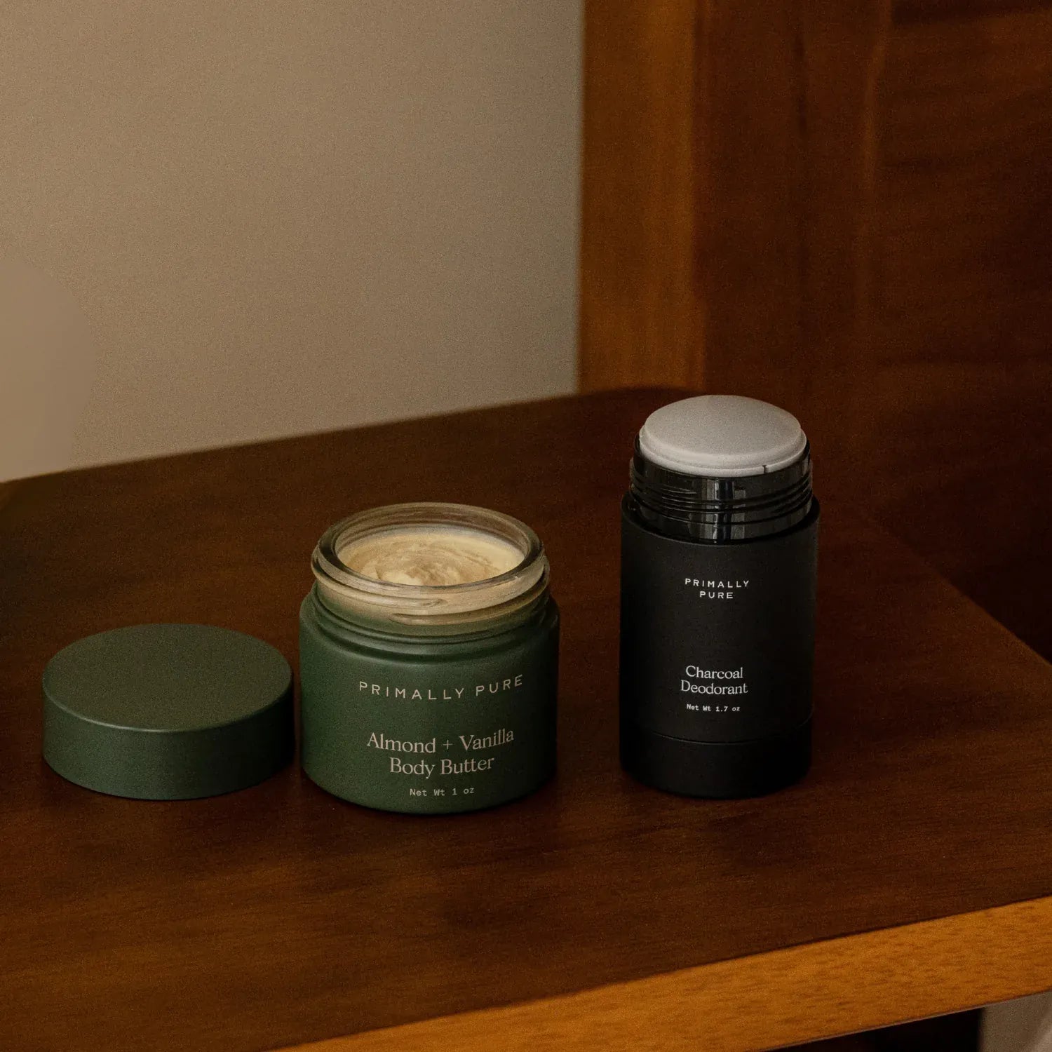 Primally Pure Everyday Body Duo Bundle displayed on a wooden table, featuring an open green jar of Almond + Vanilla Body Butter next to Charcoal Deodorant.