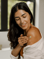 Smiling woman applying Primally Pure Everything Balm to her shoulder in a well-lit room.