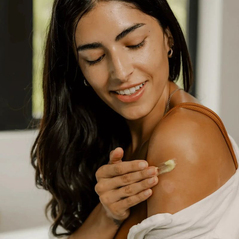 Smiling woman applying Primally Pure Everything Balm to her shoulder in a well-lit room.