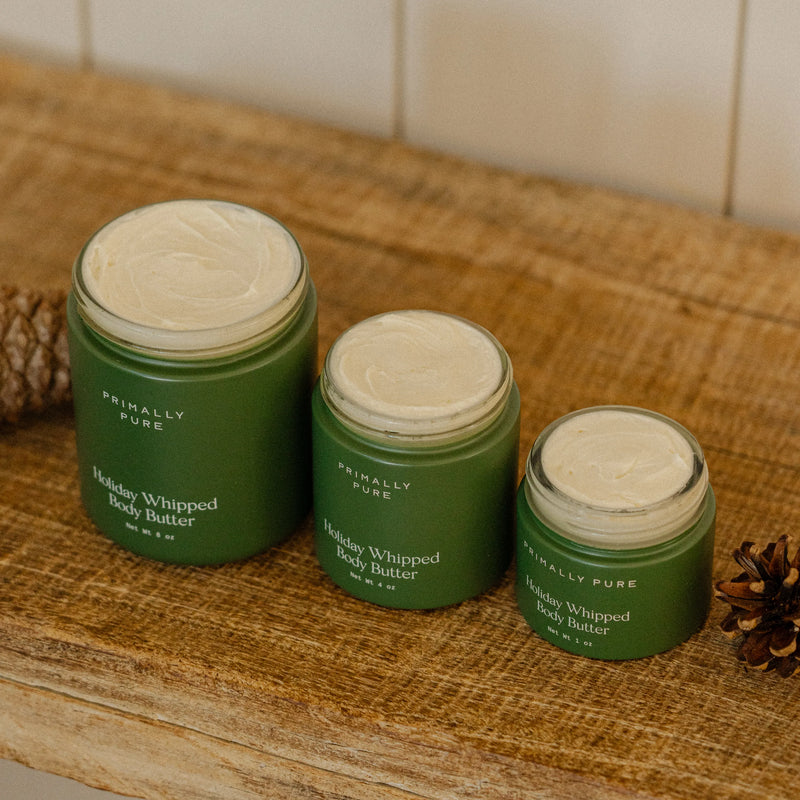 Three open jars of Primally Pure Holiday Whipped Body Butter in different sizes, arranged on a rustic wooden surface with pinecones.