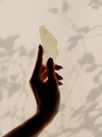 Hand holding a jade gua sha tool against a light background with leaf shadows.