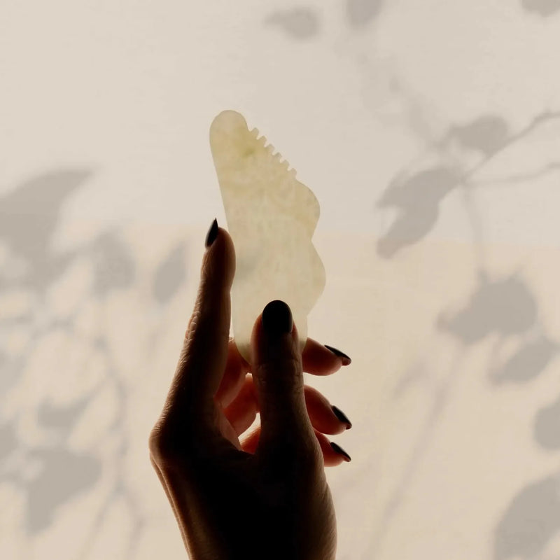 Hand holding a jade gua sha tool against a light background with leaf shadows.
