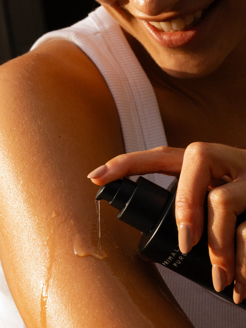 A close-up lifestyle shot of a woman applying a hydrating, Jasmine body oil onto her arm. The warm sunlight highlights the smooth, moisturizing texture of the oil on her skin, and she is wearing a white ribbed tank top.