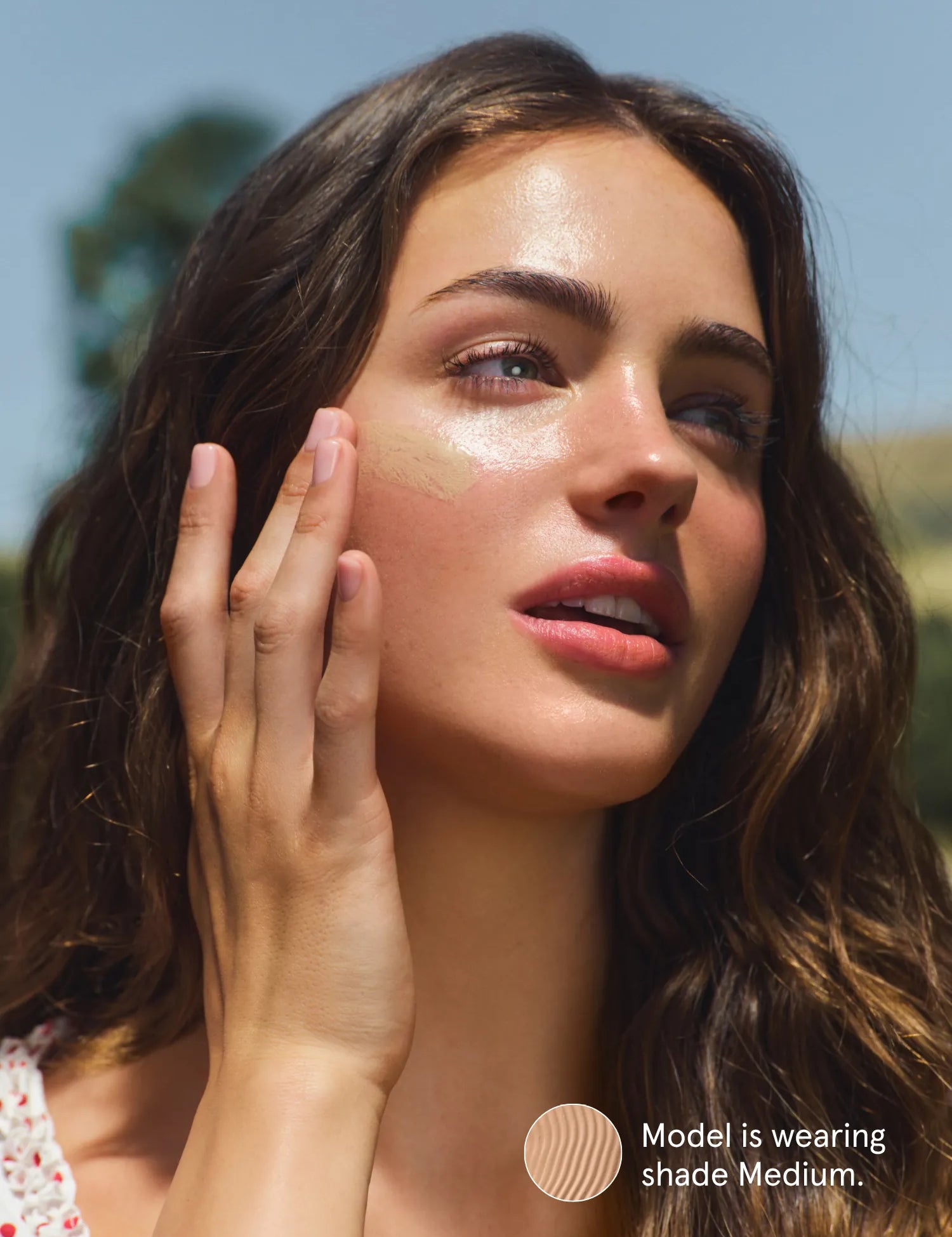 Woman applying Primally Pure Sun Serum Tinted SPF 30 in shade Medium to her face with a clear sky in the background