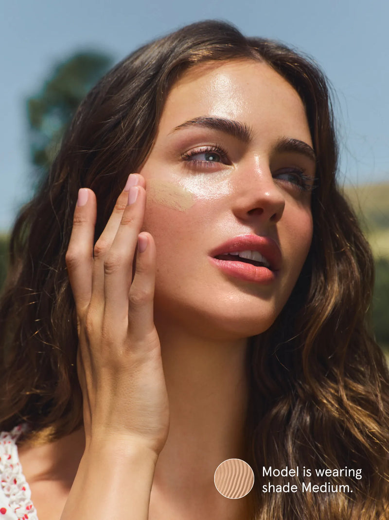 Woman applying Primally Pure Sun Serum Tinted SPF 30 in shade Medium to her face with a clear sky in the background