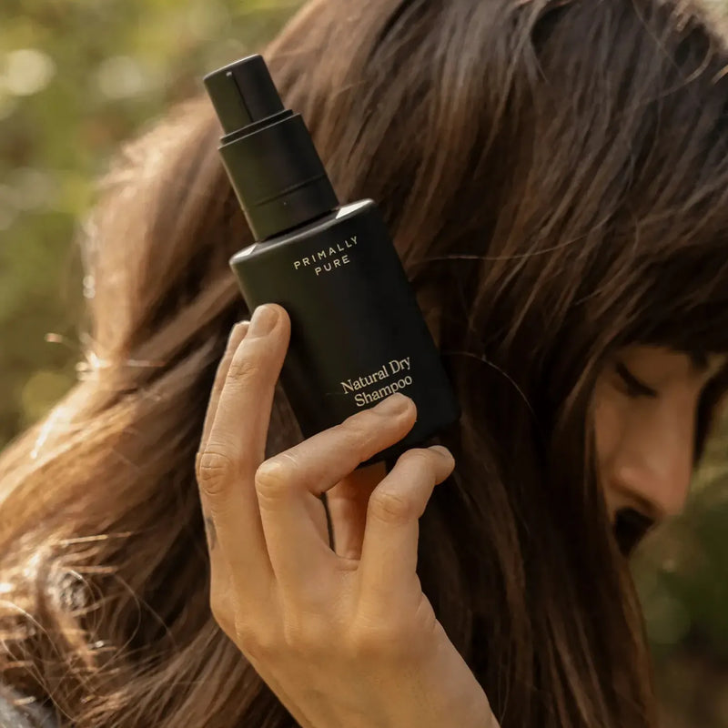 Person holding Primally Pure Natural Dry Shampoo in a black bottle, designed for refreshing hair naturally.