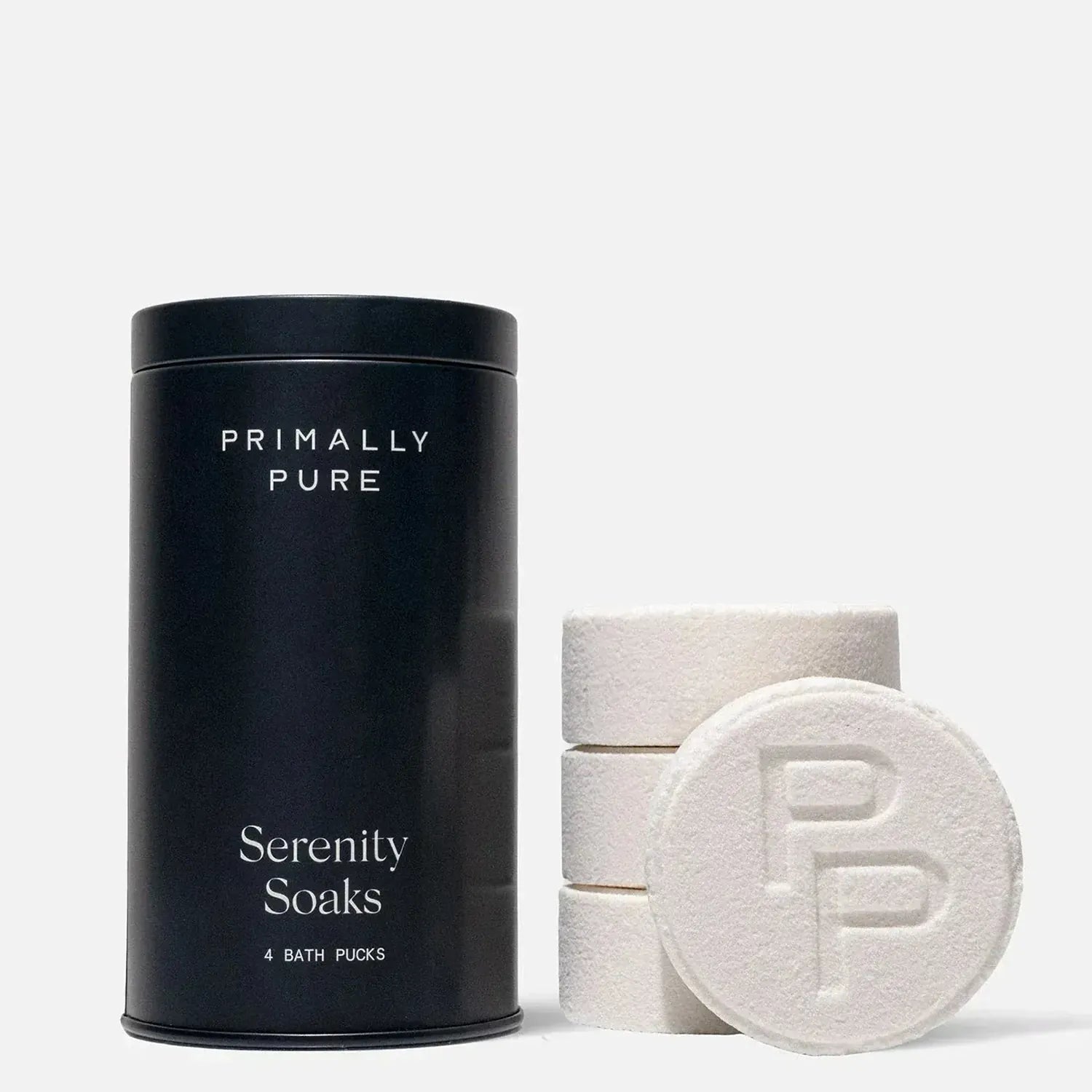 Primally Pure Serenity Soaks bath pucks shown with a matte black cylindrical tin and four round white bath pucks, including one in front with an embossed PP logo, on a light gray background.