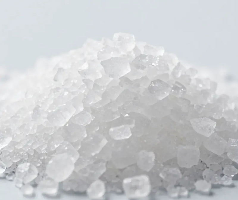A close-up pile of translucent white coarse crystals or salt grains on a light gray background.