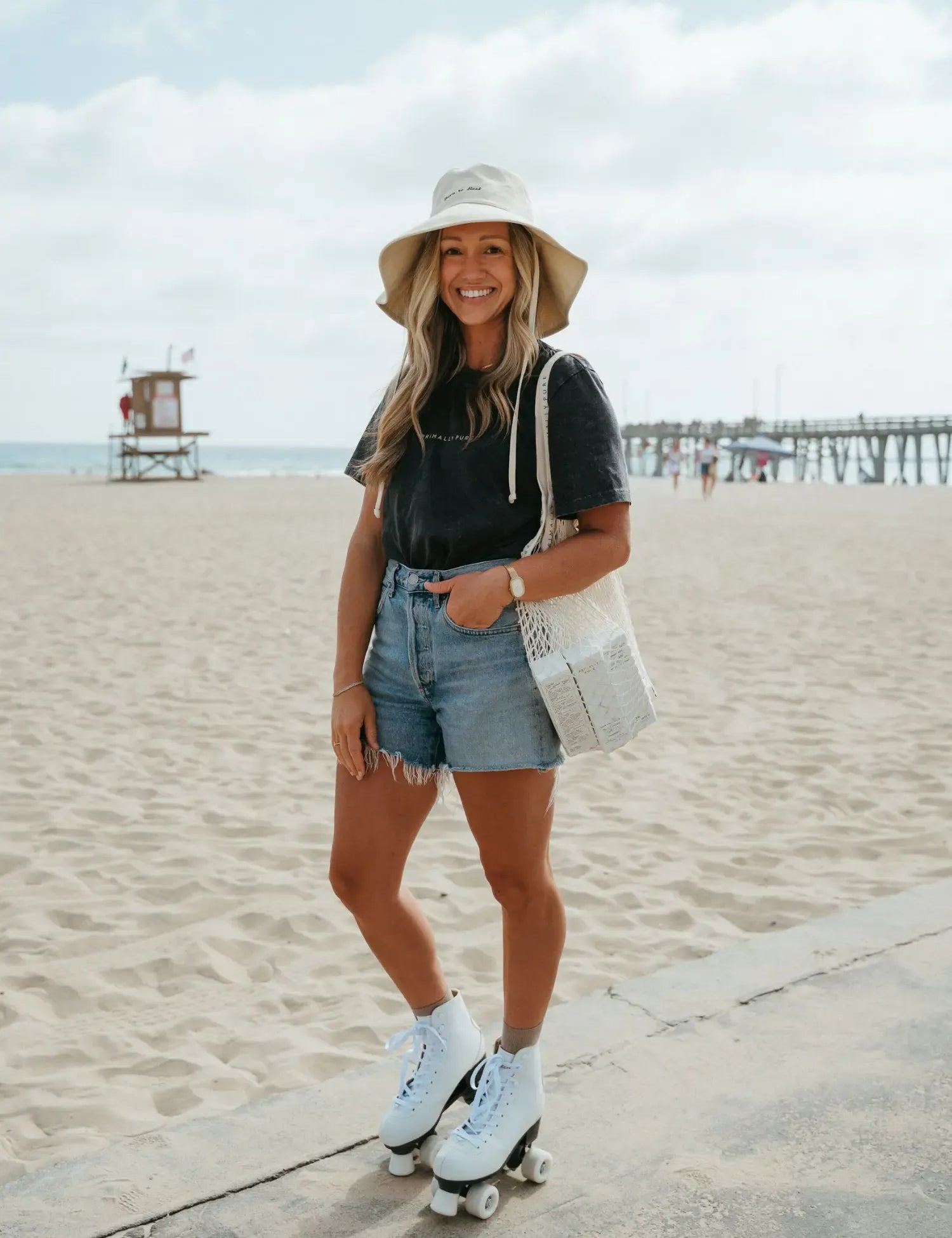 A woman posing on a beach with roller skates, wearing a beige bucket hat, a black graphic t-shirt, denim shorts, white socks, and light blue roller skates, while carrying a white woven tote bag.