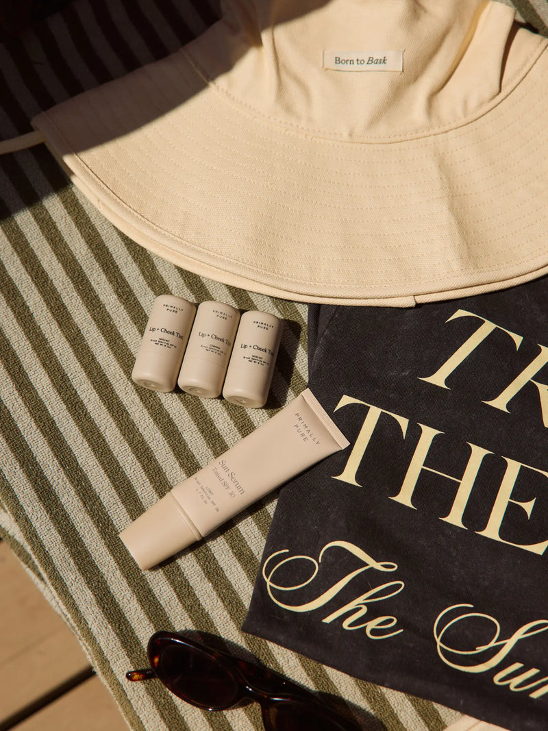 A styled flat-lay featuring a cream-colored bucket hat, a black towel with gold lettering, three small skincare tubes, a cream tube, and sunglasses on a green and white striped blanket.