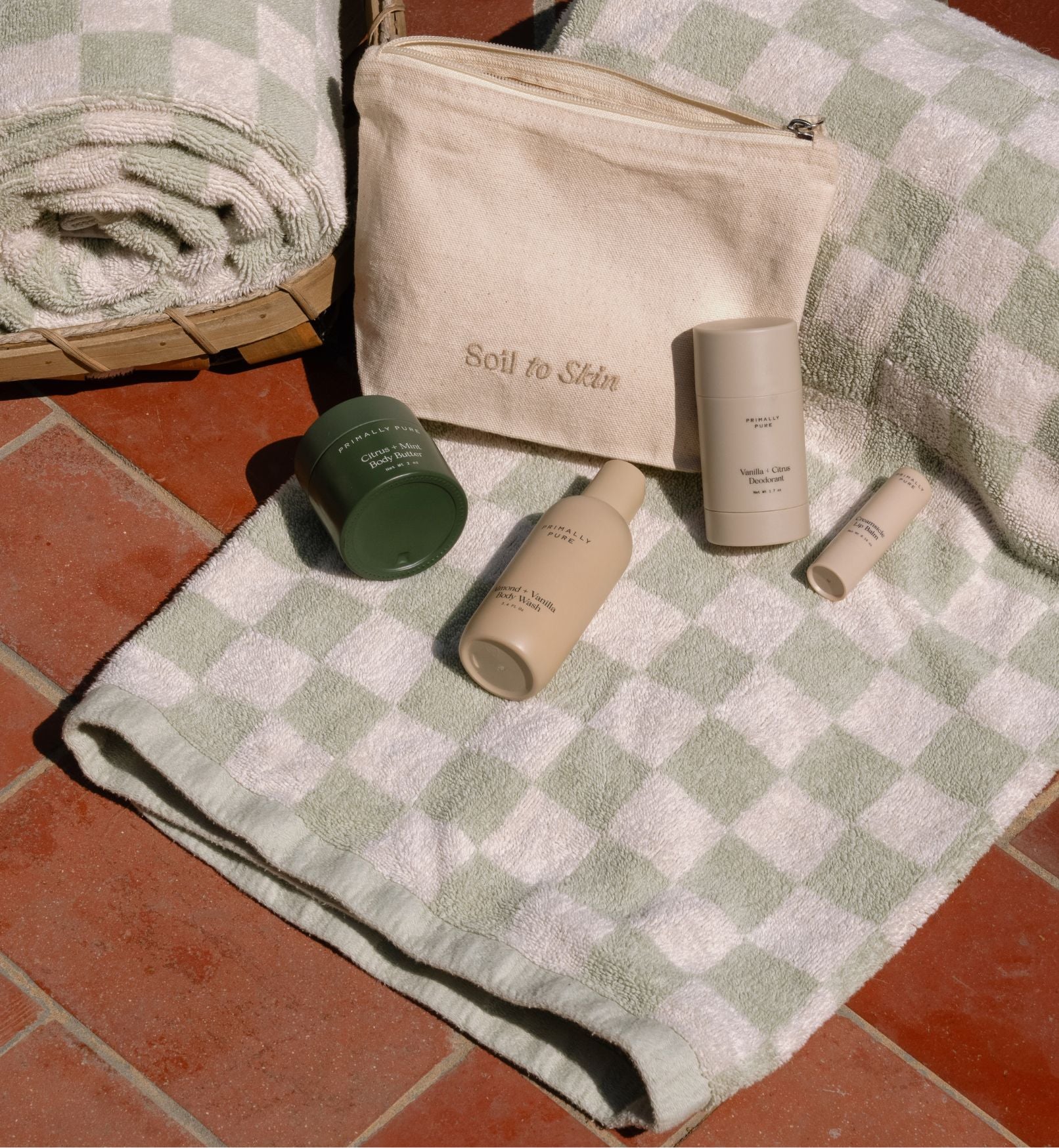 Primally Pure Cosmetic bag and travel sized skincare products on a checkered towel with 'Soil to Skin' tote branding.