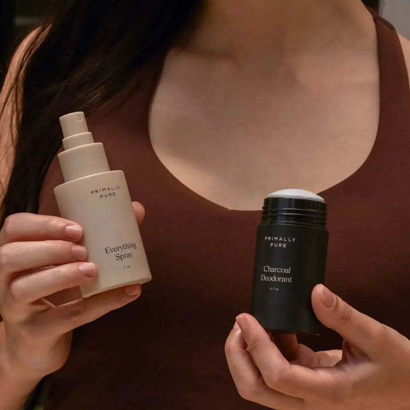 A person in a brown tank top holds two Primally Pure products:  Everything Spray bottle in one hand and a Charcoal Deodorant stick in the other.