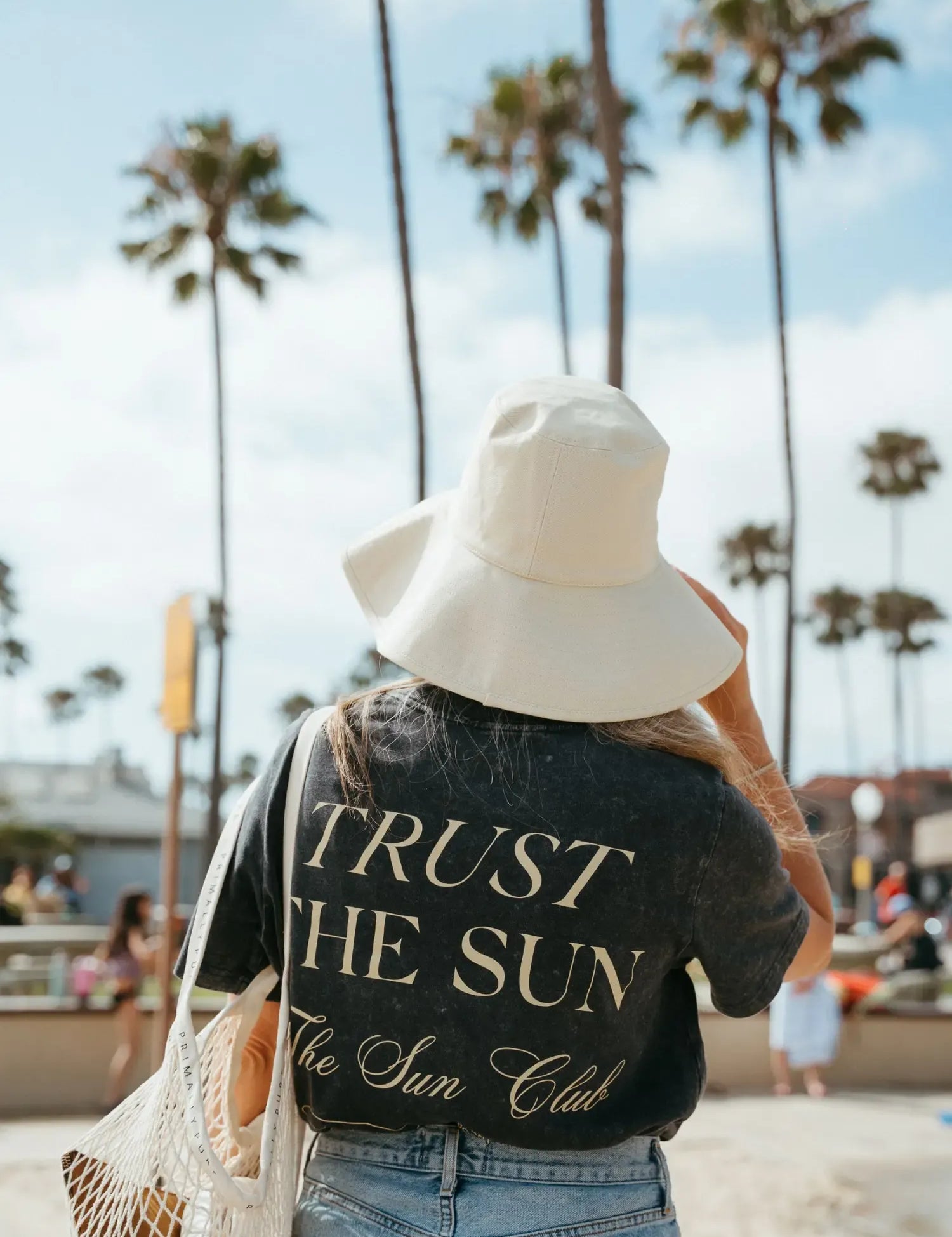 A woman viewed from behind on a beach wearing a black graphic t-shirt with 'TRUST THE SUN The Sun Club' printed in gold, a large white sun hat, and blue jeans. Palm trees and a sandy boardwalk are visible in the background.