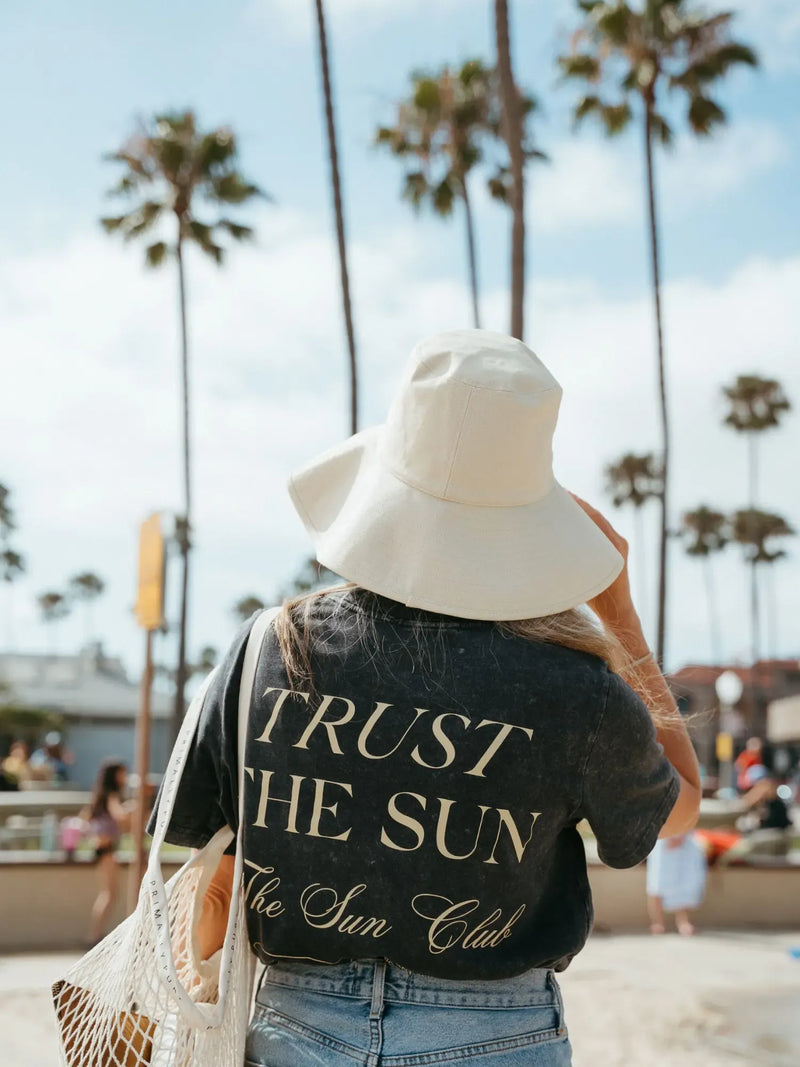 A woman viewed from behind on a beach wearing a black graphic t-shirt with 'TRUST THE SUN The Sun Club' printed in gold, a large white sun hat, and blue jeans. Palm trees and a sandy boardwalk are visible in the background.