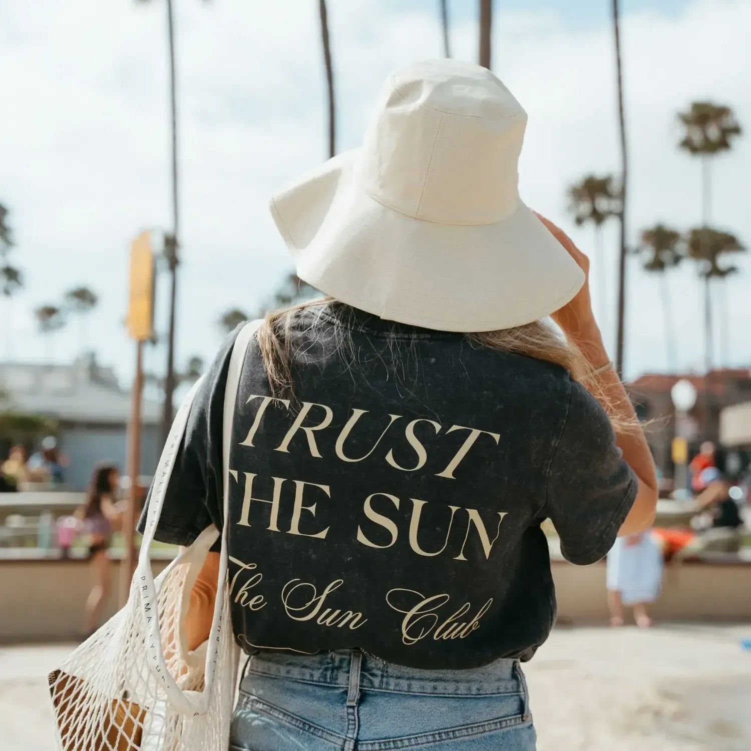 A woman seen from behind wearing a black graphic t-shirt with 'TRUST THE SUN The Sun Club' printed in beige, a large white bucket hat, and carrying a white mesh tote bag on a beach with palm trees in the background.