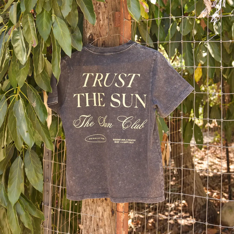 A dark gray acid-wash t-shirt hanging on a wooden fence post outdoors, featuring the text 'TRUST THE SUN The Sun Club' in cream lettering.