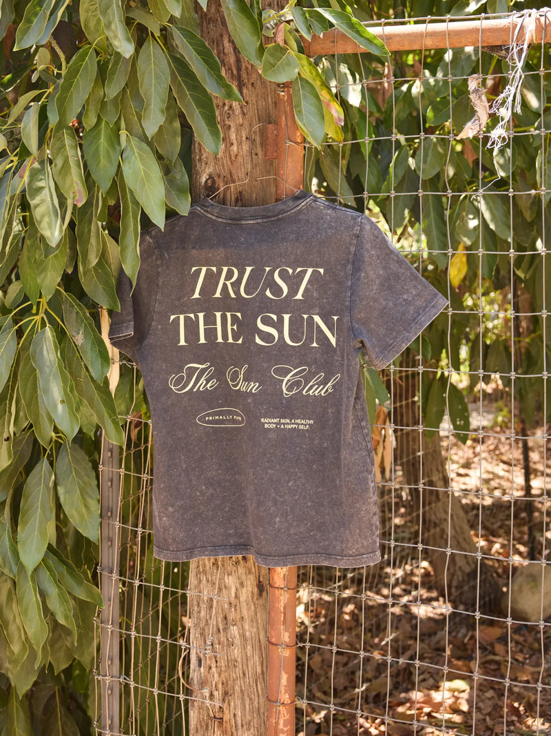 A dark gray acid-wash graphic t-shirt hanging on a wooden fence post outdoors, featuring the text 'TRUST THE SUN The Sun Club' in cream lettering.