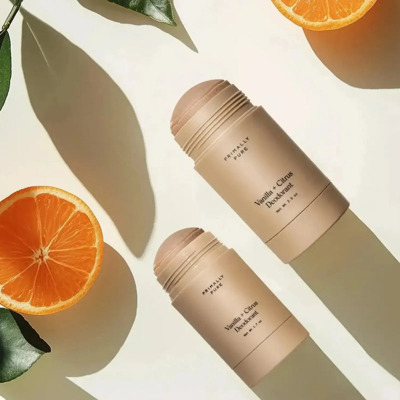 Two Vanilla + Citrus Primally PureDeodorant stick containers arranged diagonally on a warm cream background with stylized orange slices and leaves casting shadows.