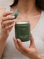 Woman holding a jar of Primally Pure Almond + Vanilla Body Butter with the lid open.